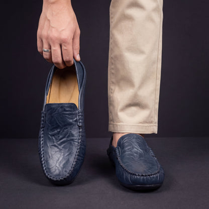 Een man draagt de marineblauwe Jorin leren pantoffels, gecombineerd met een casual broek, zittend op een houten vloer.