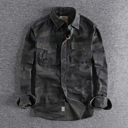 Mannelijk model draagt een camouflage T-shirt in olijfgroen, staand in een stedelijke omgeving met een ontspannen houding.