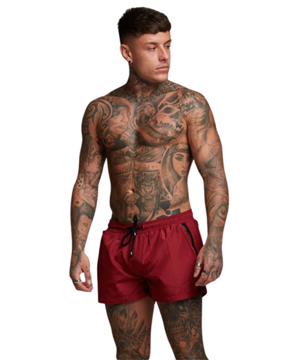 Max zwemshort voor heren in rode kleur, vooraanzicht, liggend op een zonnig strand met zand en water op de achtergrond.