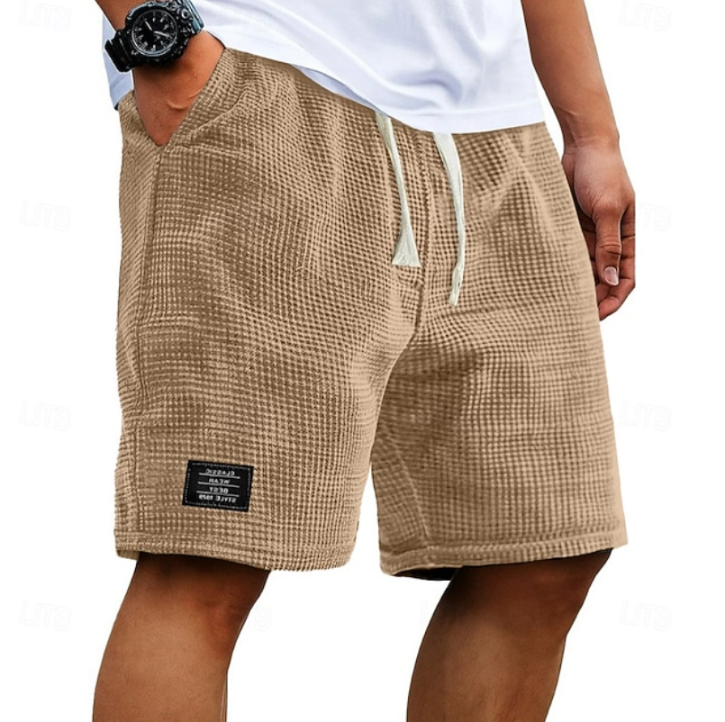 Een man draagt de Nils wafelkatoenen shorts in beige, gecombineerd met een licht T-shirt, ontspannen zittend.