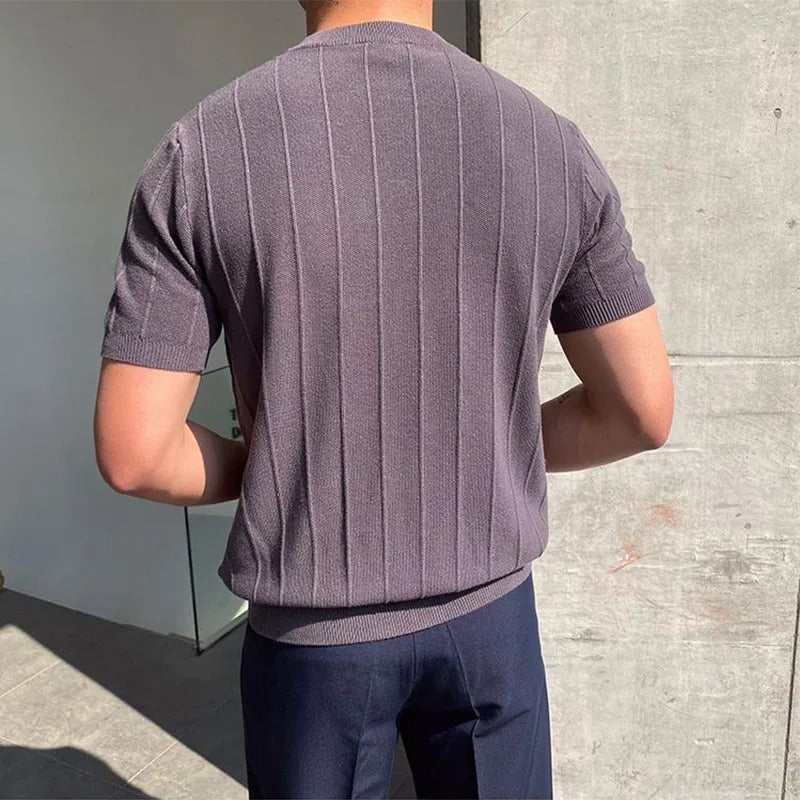 Een man in een donkergrijs Reuben T-shirt in een stedelijke omgeving, straalt zelfvertrouwen uit.