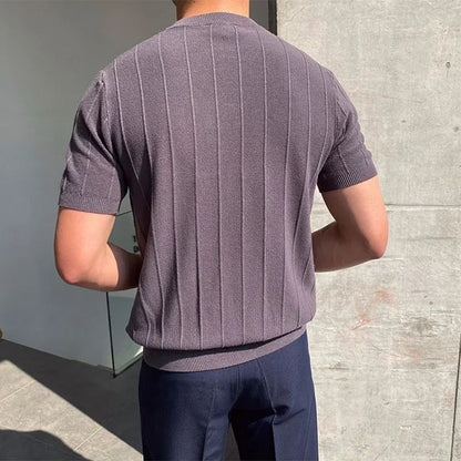 Een man in een donkergrijs Reuben T-shirt in een stedelijke omgeving, straalt zelfvertrouwen uit.