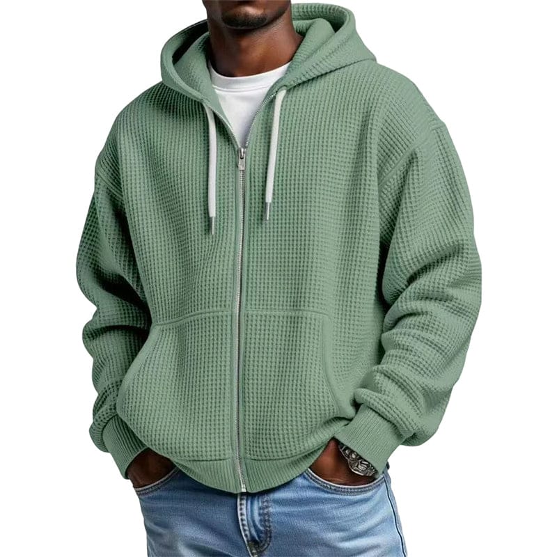 Man in een lichte groene wafeltextuur hoodie met rits, staand en glimlachend, in een buitenomgeving.